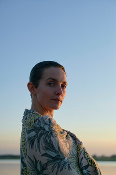 A woman with her hair pulled back stands in the evening light, looking over her shoulder with a calm expression against a clear sky.