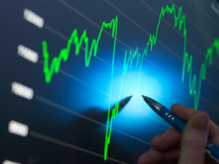 A person's hand holds a pen, pointing at a bright screen displaying a stock market graph with upward trends in green and blue tones.