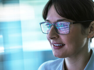 A smiling woman with glasses, the reflection of a screen visible on her lenses, against a softly lit blue background.