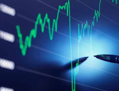 A close-up of a stock market graph on a digital screen with a person's hand holding a pen pointing to a specific trend line.