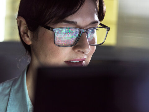 A focused individual wearing glasses with computer code reflection in the lenses is intently looking at a computer screen.