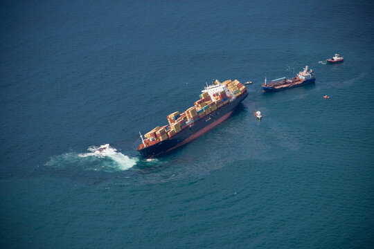 An aerial view of a large cargo ship in trouble at sea, being accompanied by several smaller vessels.