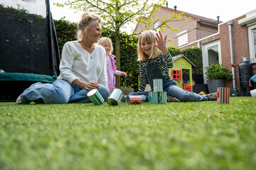 An elderly woman and two young girls are sitting on a grass lawn playing with colorful tin cans, set up like bowling pins, with a child's playhouse in the background.