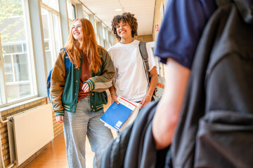 Two students are walking down a school hallway, smiling and engaging in conversation, with books in hand.
