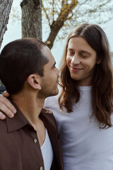 Obraz premium A gay couple in casual attire stands joyfully together, surrounded by lush greenery, next to a towering tree.