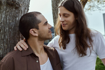 a loving gay couple, stand together near a tree in casual clothes, enjoying quality time outdoors.