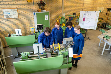 Four individuals in blue work attire are operating green industrial machinery in a workshop setting, collaborating on a technical task.