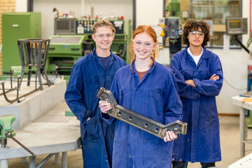 Three smiling teenagers in protective blue work overalls stand confidently in a workshop environment, with one holding a mechanical part.