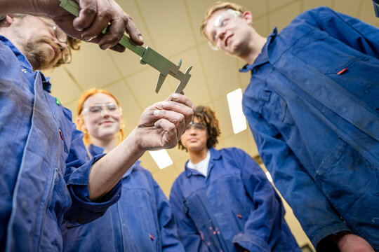 A group of four individuals in blue lab coats are focused on an object being measured with a caliper by one of them, likely in an educational or engineering setting.