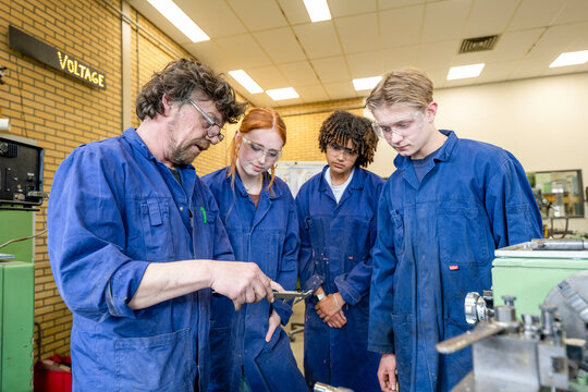 A group of students wearing blue coveralls attentively observes a demonstration by an instructor in a technical workshop environment.