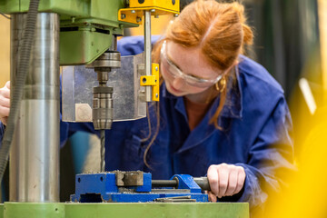 A red-haired woman in safety glasses operates a drilling machine in an industrial setting.