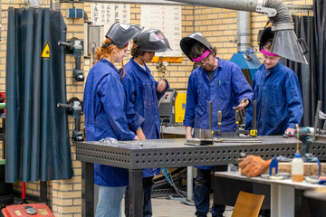 Four people dressed in protective work gear, including welding helmets, are engaging in a welding activity at a workshop table equipped with metalworking tools.