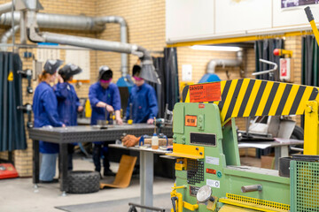 Workers in blue uniforms are focused on their tasks at different stations in an industrial workshop setting, with machinery and safety warnings visible.