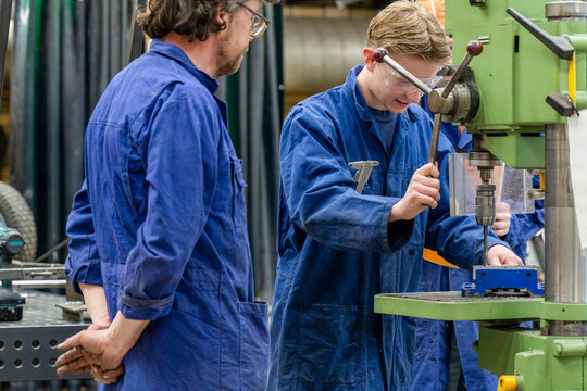 Two workers in blue overalls are operating machinery in an industrial setting, with one observing while the other concentrates on the task.