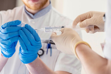 A healthcare professional is administering an injection to a patient, who is out of focus in the background, while wearing blue and yellow gloves.