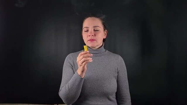 A charming young woman exhales a thick cloud of vapor, e-cigarette. With a casual, unbothered expression set on the stark black backdrop, youthful indulgence and the cavalier embrace of trending habit