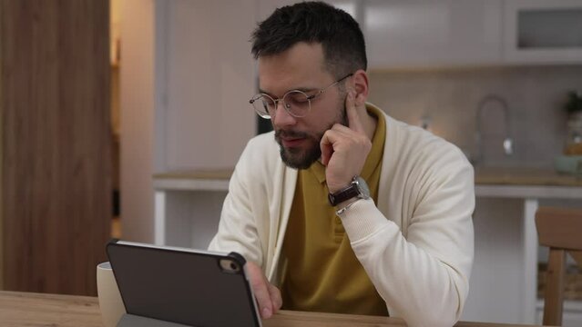 Adult caucasian man sit at kitchen look at tablet and have headache