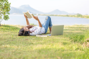 Girl enjoy listening music and reading a book and play laptop on the grass field of the park in the...