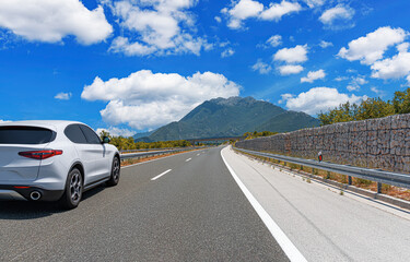 A white car drives along the highway against the backdrop of rocky mountains on a sunny day.