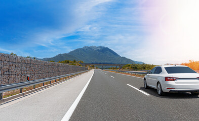 A white car drives along the highway against the backdrop of rocky mountains on a sunny day.