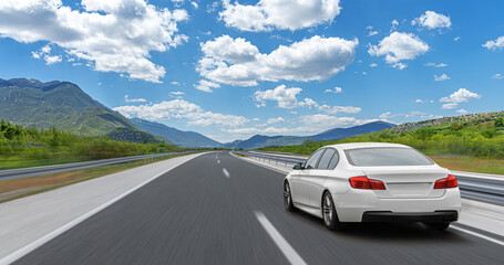 A white car drives along the highway against the backdrop of rocky mountains on a sunny day.