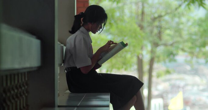 An Asian high school student girl in white uniform sits and read a book anxiously to prepare for exams in the evening after school while waiting for her parent to take her home.