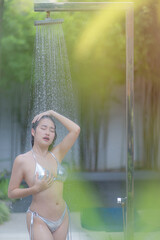 Beautiful young Asian woman in a silver color bikini takes a shower after swimming in the ocean, sea, or pool, relaxing, feeling fresh, and cooling on vacation holiday.