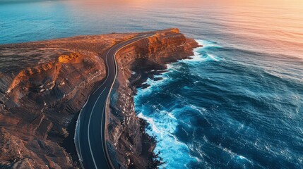Aerial view of a curving asphalt road hugging a rugged coastline, with deep blue ocean waves crashing against the cliffs at sunset. - Powered by Adobe