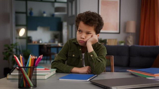 Sad upset tired negative offended frown boring little ethnic multiracial multiethnic American African boy son kid child schoolboy pupil at home education studying distant at table learning study alone