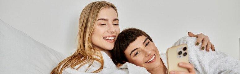 A beautiful lesbian couple in bath robes lay snuggled under a cozy blanket on a bed in a hotel room.