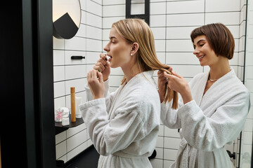 A young lesbian couple in bath robes, one flossing teeth, other braiding hair in a hotel bathroom.