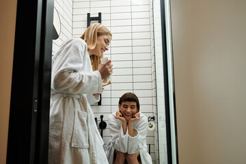 A woman in a bathrobe brushes her teeth while her partner watches in a hotel bathroom.