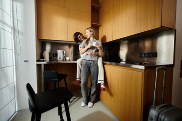 A man tenderly holds a woman in a warm kitchen, both enjoying coffee cups in a hotel room setting.