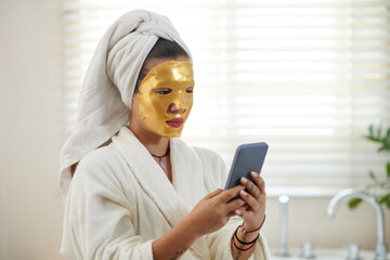 Portrait of woman with golden sheet mask on her face texting friends