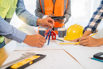 An architect engineer meeting is taking place at a desk covered with blueprints. They are discussing plans for a house, an apartment, and a condo, focusing on design and renovation strategies.