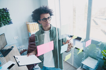 Photo of cheerful handsome agent dressed khaki shirt eyewear reading documents creating plan indoors workshop workstation