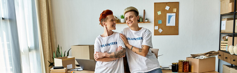 Two young women in volunteer t-shirts standing side by side in a room, united in their charitable...