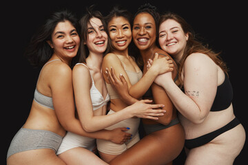 Diverse group of five women dressed in underwear embrace each other and smiling warmly against...