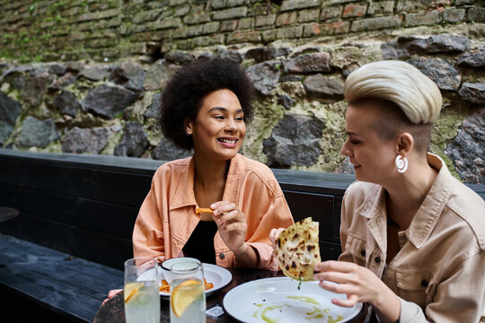 Two diverse, beautiful women sit at a table, happily eating pizza in a cozy cafe.