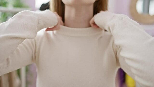Close-up of a thoughtful caucasian woman adjusting her beige sweater indoors, in a room with colorful wardrobe.