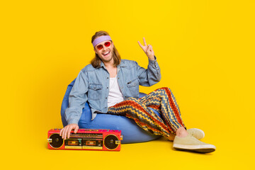 Full body portrait of cool young man sit bag show v-sign wear denim shirt isolated on yellow color...