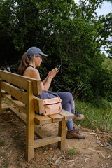 Girl in her forties sends messages sitting on a bench, resting from a hiking route with her backpack.