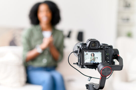 An African-American woman with a radiant smile is seated on a couch, interacting with a camera on a gimbal, creating content in a modern living room with soft natural light, focus on the camera