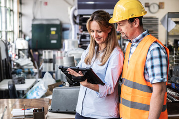 A supervisor stands confidently with a worker on the factory line, using a tablet to check the...