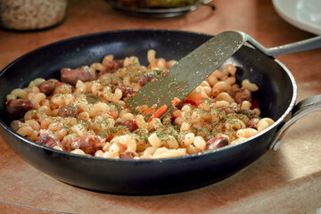 Spiral pasta in sauce with chicken hearts, vegetables and dill in a skillet on a kitchen table with plastic spatula