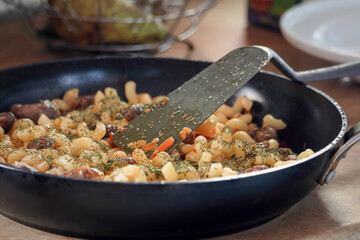 Spiral pasta in sauce with chicken hearts, vegetables and dill in a skillet on a kitchen table with plastic spatula close up