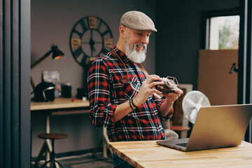 Portrait d'un homme photographe hipster situé dans un studio atelier et qui tient un appareil photo vintage en souriant devant un ordinateur