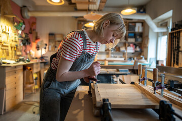 Concentrated female carpenter working on gluing wooden planks together, using clamping brackets and bar clamps. Woodworking in carpentry workshop. Small family furniture manufacture. 