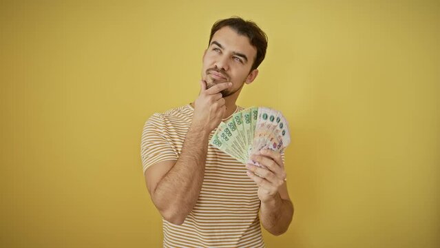 Thoughtful young hispanic man, holding money with a serious face, hand placed on chin, thinking about a questionable idea, isolated on a vibrant yellow background.