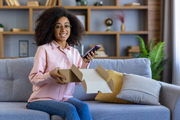 Obraz premium Smiling woman unboxing a package and using her smartphone while relaxing on a couch in a cozy living room.